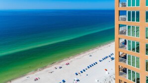 A beach with clear blue water, a sandy shore, and a building with balconies overlooking the beach. - Phoenix Gulf Shores 803 3 Bedroom Condo by RedAwning (Gulf Shores)