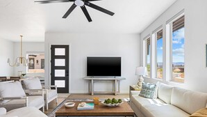 A modern living room with a flat-screen TV, a ceiling fan, and large windows offering a view of the outdoors. - Beachcomber Bliss 8 Bedroom Townhouse (St. George)