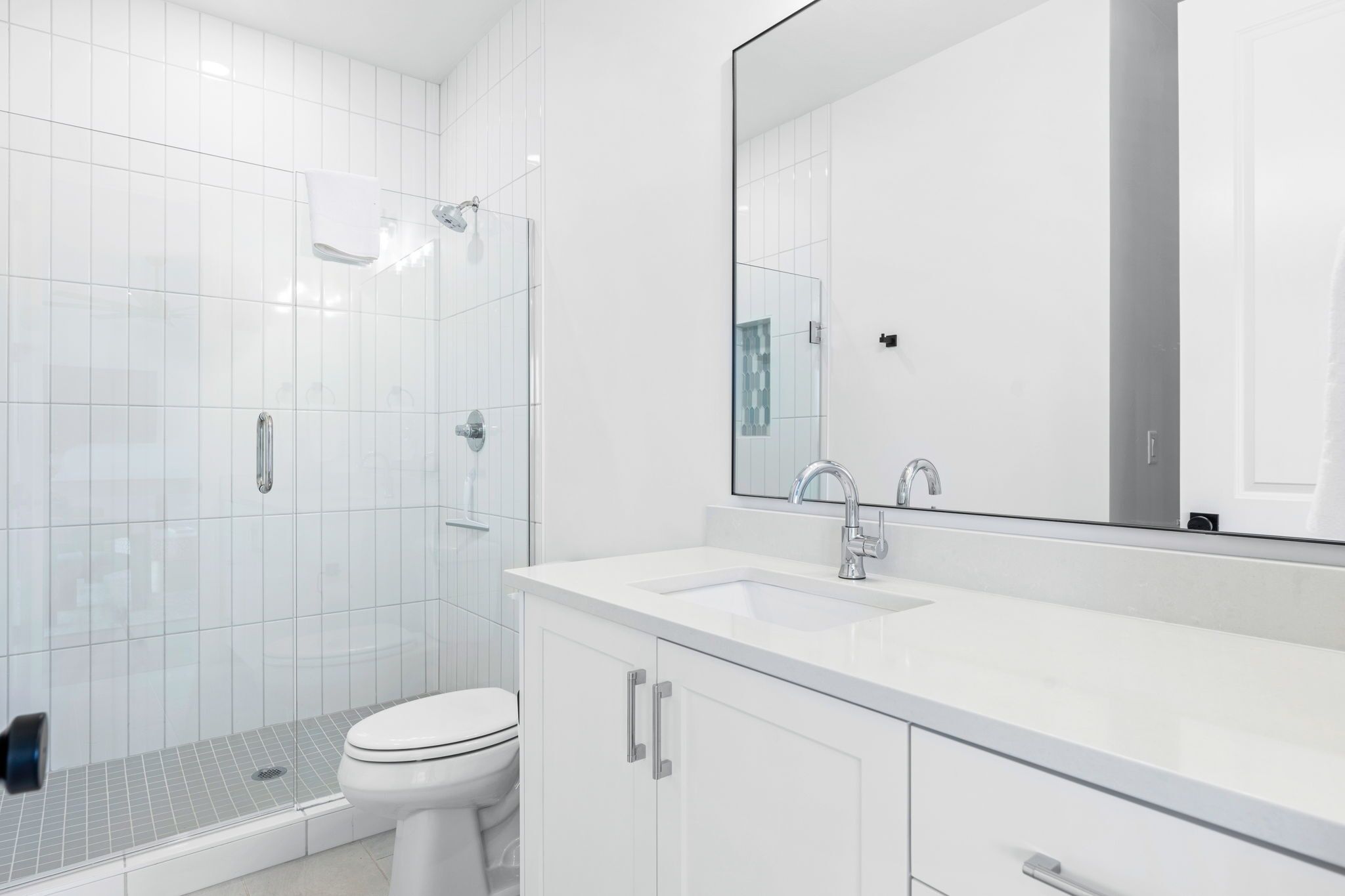 Un bany modern amb una cabina de dutxa de vidre, un vàter blanc i un mirall gran sobre un lavabo blanc.