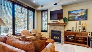 A living room with a fireplace, a TV mounted above it, and a painting on the wall. - Woodwinds #1303 4 Bedroom Condo by RedAwning (Mammoth Lakes)