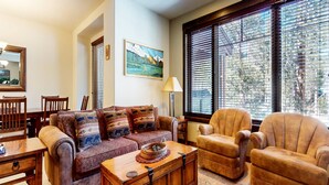 A living room with a sofa, armchair, and a coffee table. There is a painting on the wall, a lamp, and a window with blinds. - Woodwinds #1303 4 Bedroom Condo by RedAwning (Mammoth Lakes)