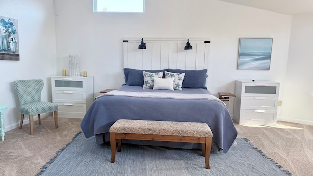 Un dormitorio con una cama, dos mesitas de noche, una silla, un banco y una ventana.