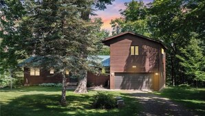 A two-story wooden cabin with a garage, surrounded by lush greenery and trees.