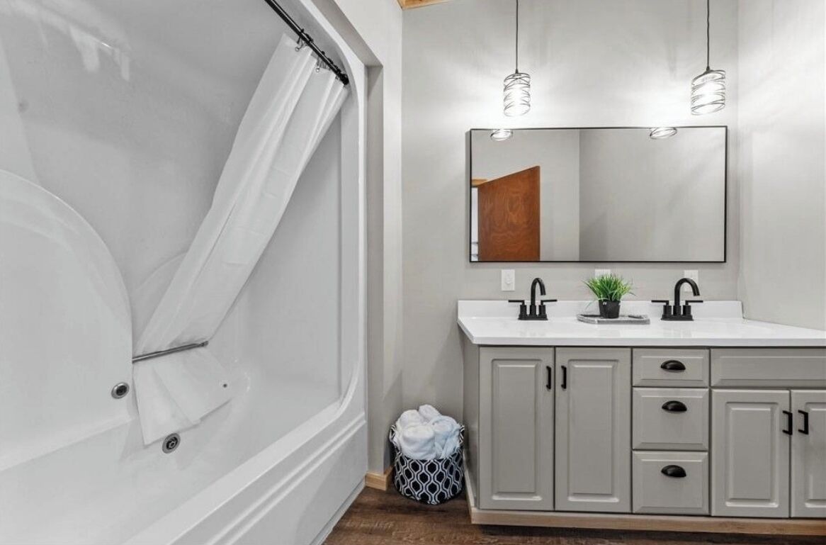 A bathroom with a bathtub, a sink with two faucets, a mirror, and a cabinet with drawers.