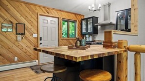 A kitchen with a wooden herringbone wall, a central island with stools, a black cabinet, and a window with greenery outside.