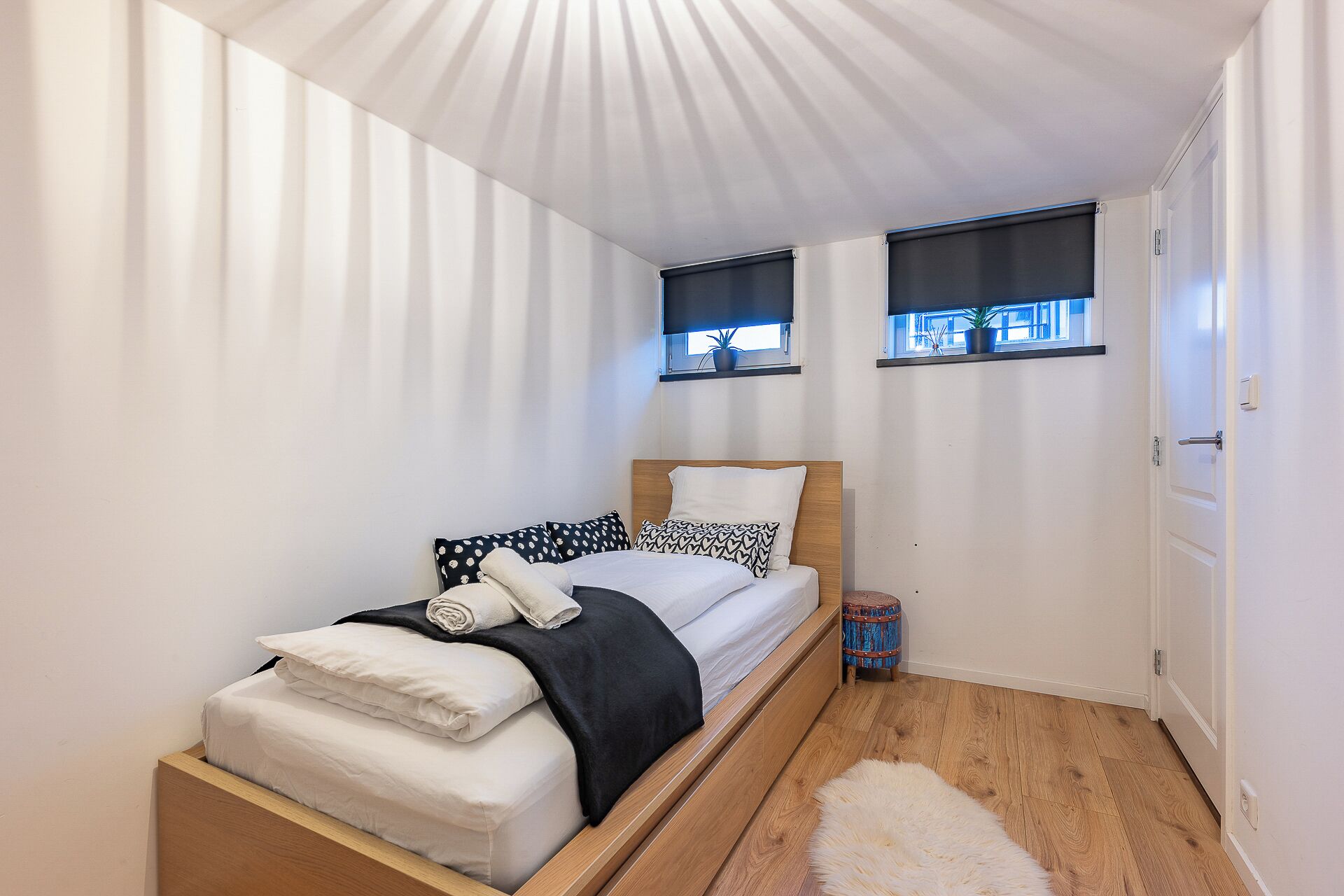 A small, neatly arranged bedroom with a single bed, two windows with blinds, a door, and a small rug.