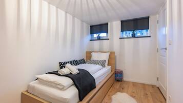A small, neatly arranged bedroom with a single bed, two windows with blinds, a door, and a small rug.