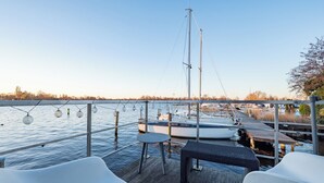 En dæk med to hvide stole, et bord og udsigt over lystbådehavnen med både og en klar himmel.