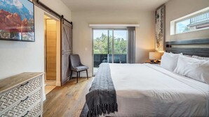 A modern hotel room with a large bed, a chair, a desk, and a balcony with a view of trees.