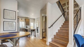 A spacious living area with a wooden staircase, a dining table with chairs, and a kitchen in the background.