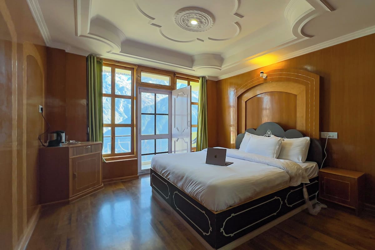 Une chambre d’hôtel avec un lit en bois, un ordinateur portable posé sur le lit, un bureau avec une bouilloire, et une vue sur les montagnes par la fenêtre.