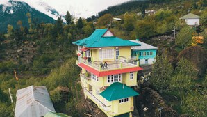 Un edificio de varios pisos con techo verde, ubicado en una zona montañosa con árboles y vistas a una cadena de montañas.