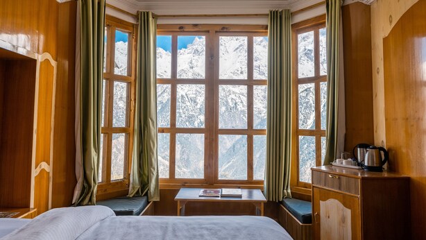 Una habitación con paredes de madera, un ventanal con cortinas, una cama, una mesita y un banco.