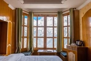 Une pièce aux murs en bois, avec une grande fenêtre ornée de rideaux, un lit, une petite table et un banc.