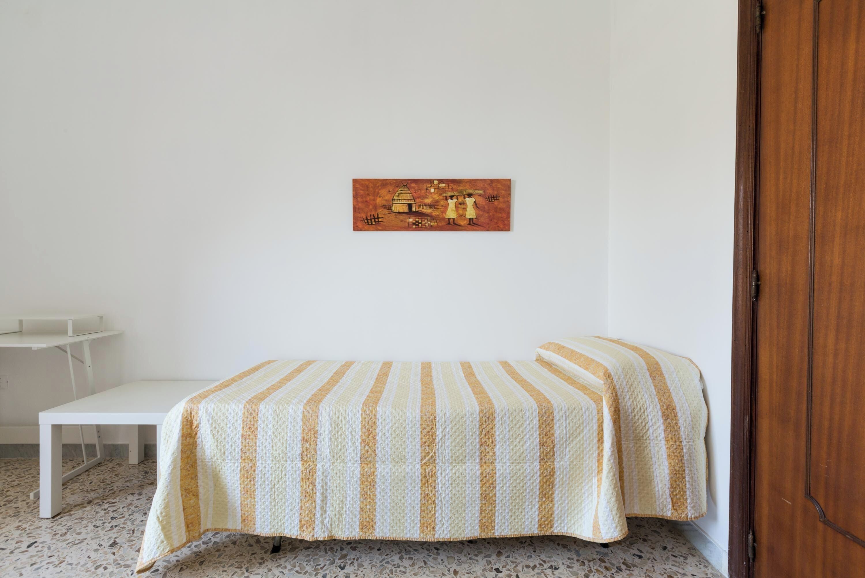 Una stanza piccola e modesta, con un letto, un tavolino e una porta.