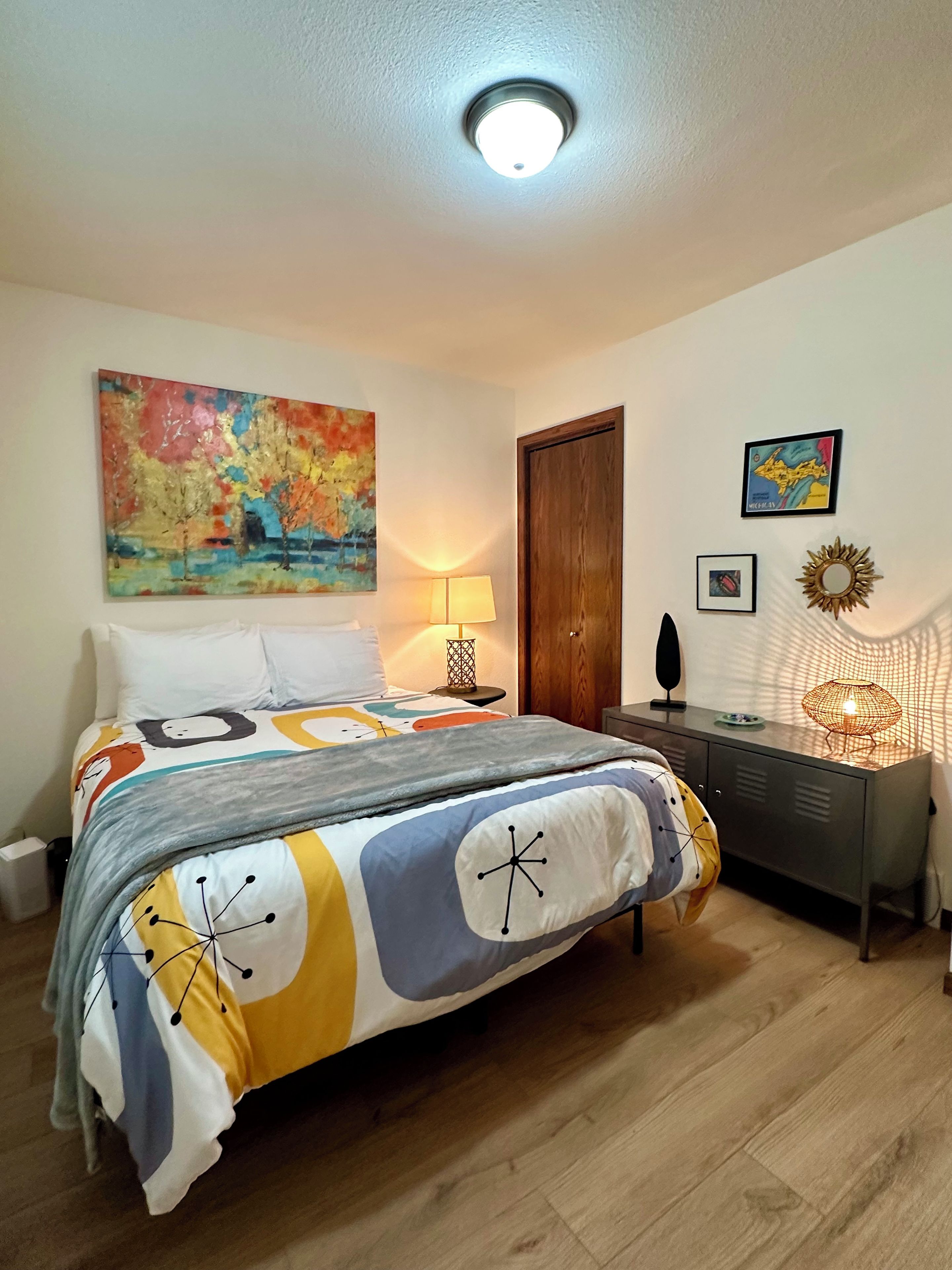 A bedroom with a bed, a nightstand, a lamp, a wall-mounted mirror, and a painting.