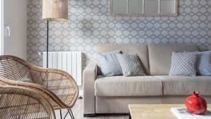 En stue med tapet med mønster, en beige sofa, en fletstol, en gulvlampe og en rød granatæble på et bord.