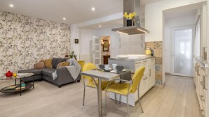 Een moderne woonkamer met een grijze bank, gele stoelen, een eettafel en een keuken met witte kasten en een grijze afzuigkap.