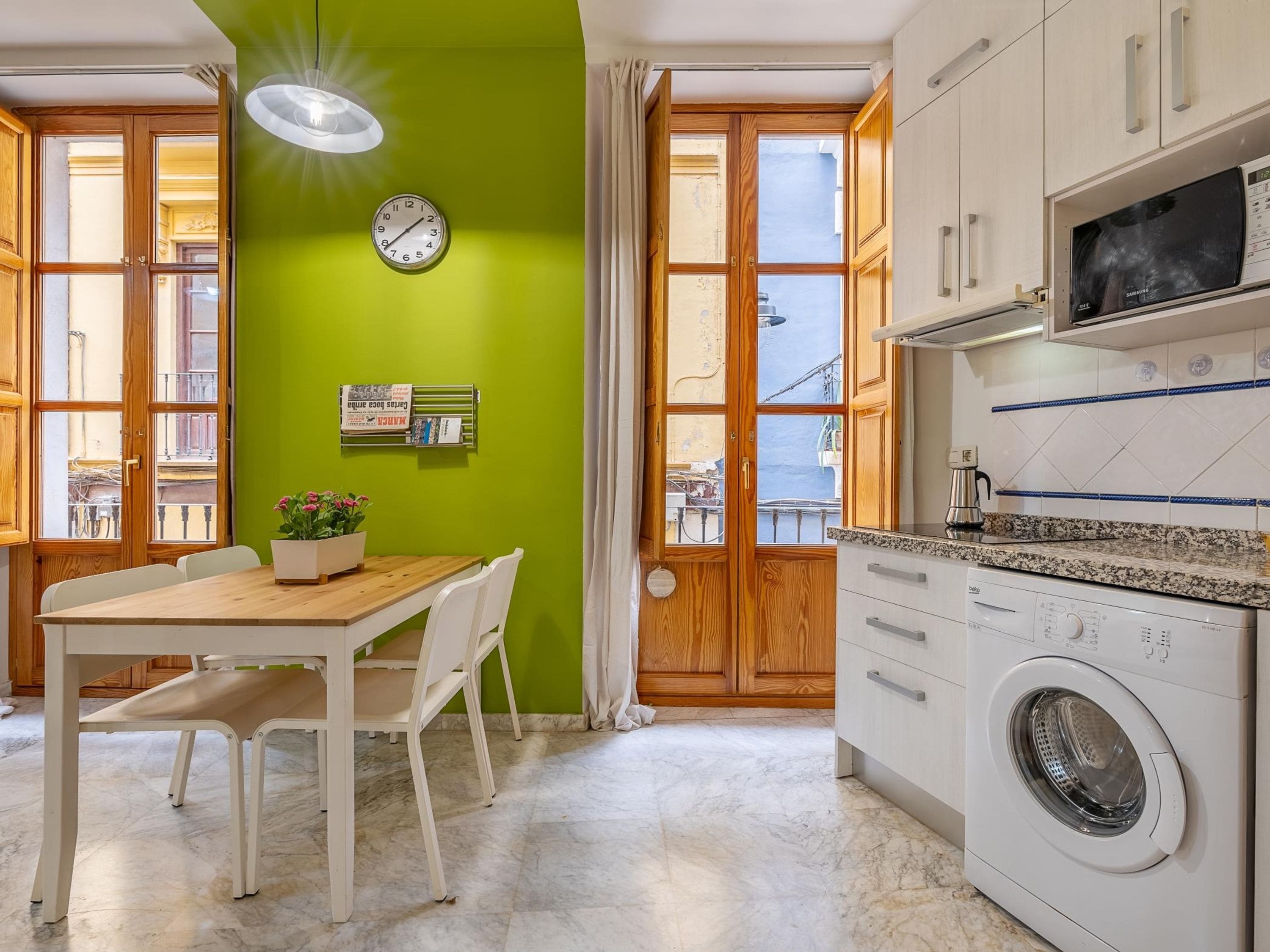 A compact kitchen with a washing machine, a clock, and a dining area with a table and chairs.
