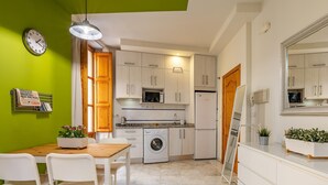 Μια μικρή κουζίνα με πλυντήριο ρούχων, φούρνο μικροκυμάτων και ψυγείο. Υπάρχει ένα τραπέζι φαγητού με καρέκλες και ένα φυτό σε γλάστρα.