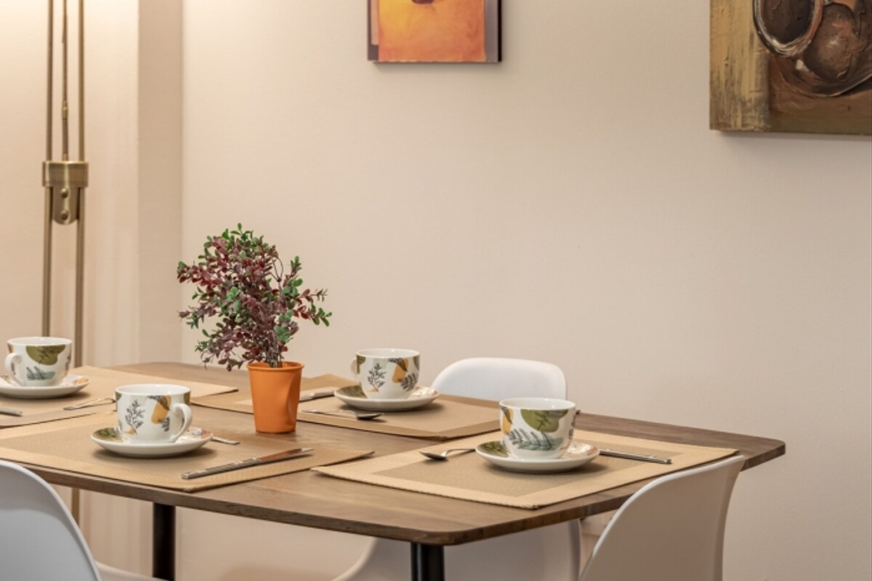 Zona pranzo con tavolo imbandito per due persone, con una pianta in vaso, due tazze e un quadro appeso al muro.