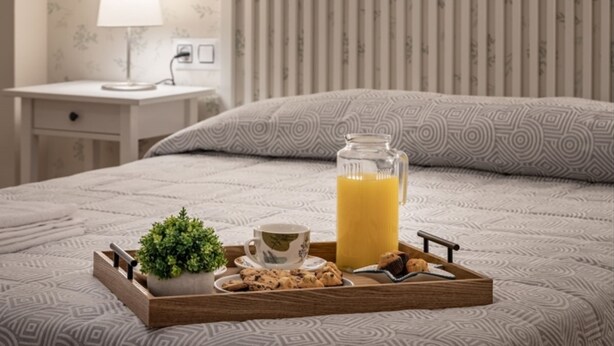 A neatly made bed with a patterned bedspread, a bedside table with a lamp, a tray with breakfast items including a glass of orange juice, a cup, cookies, and a small plant. - Luxury Suite Campo del Pr Ncipe (Granada)