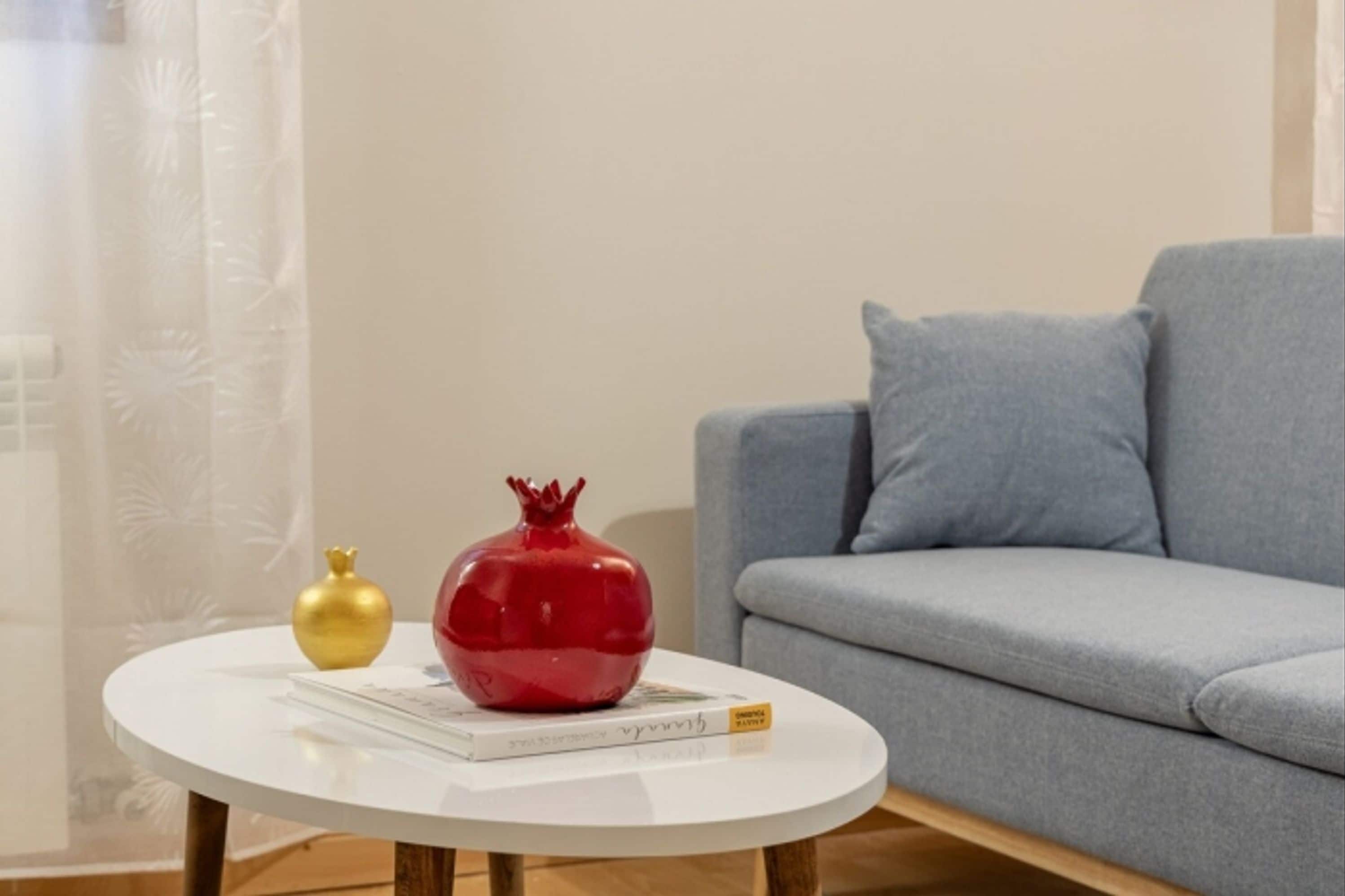 A round white table with a book and two decorative vases, a gold and a red one, in front of a grey sofa.