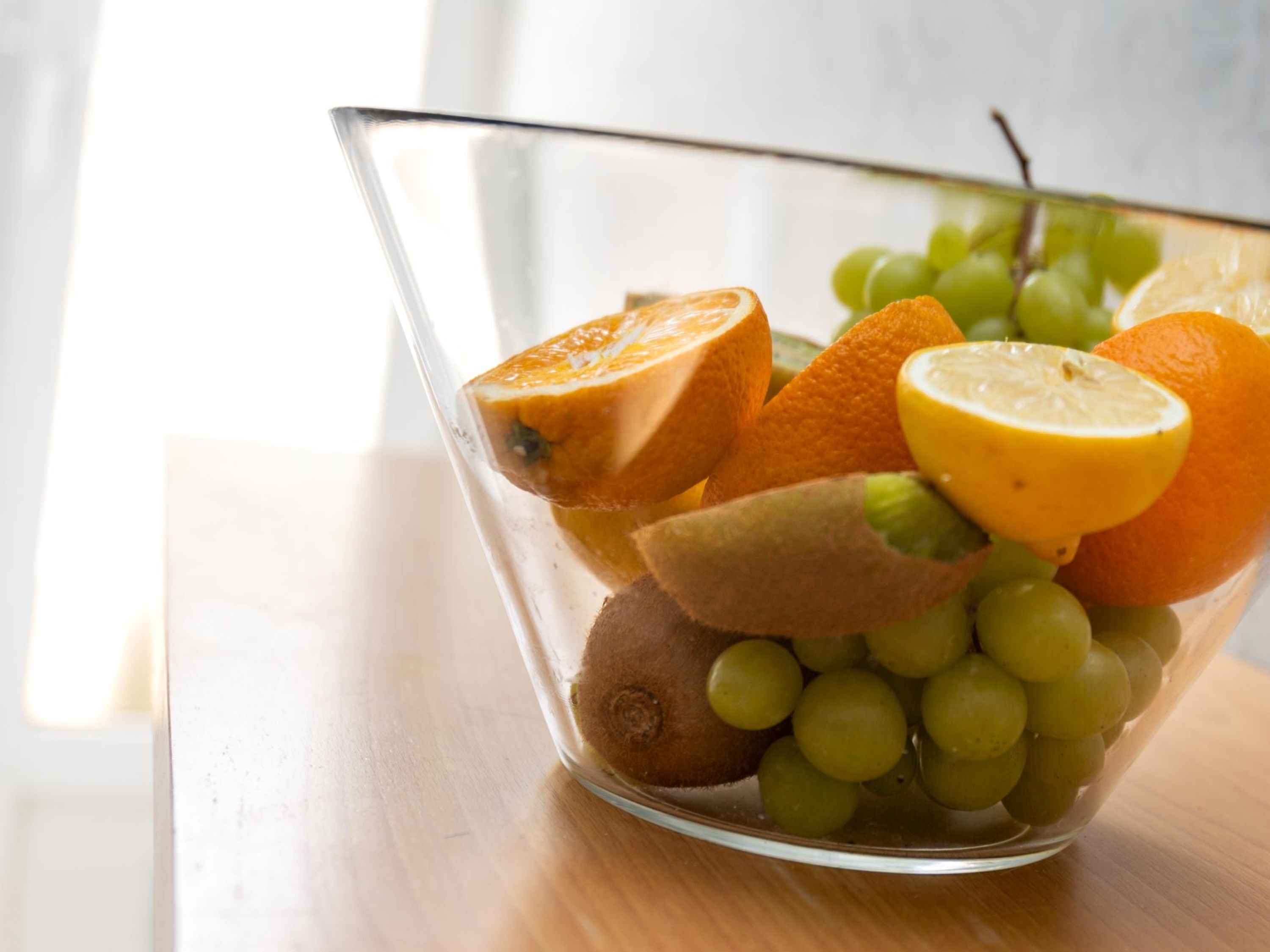 Een glazen schaal met schijfjes sinaasappels, druiven en kiwi op een houten ondergrond.