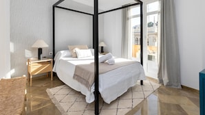 Een moderne slaapkamer met een hemelbed, nachtkastje, lamp en uitzicht op de stad door grote ramen.