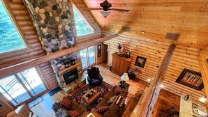 Ein geräumiges Blockhaus-Interieur mit Kamin aus Stein, Holzmöbeln und großen Fenstern.