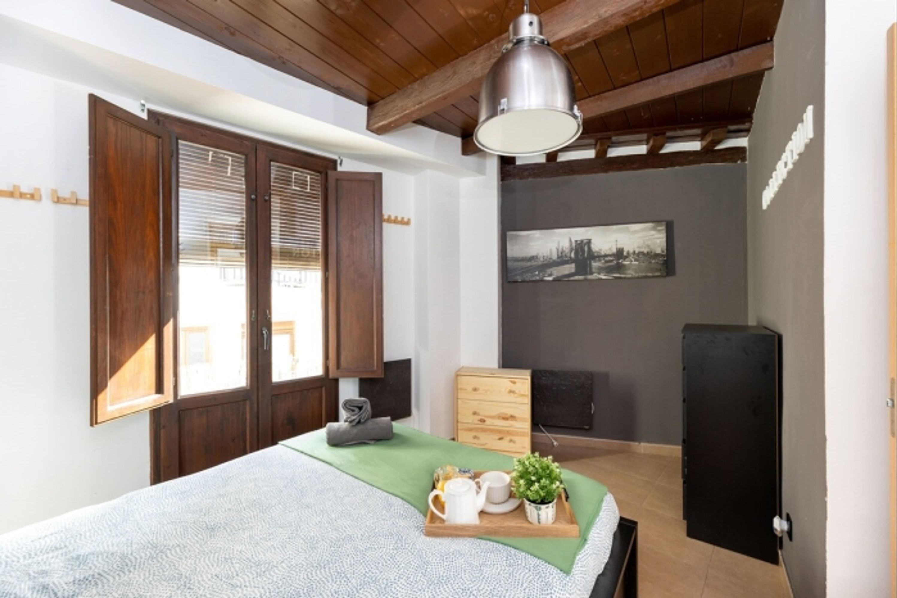 Ein Schlafzimmer mit Bett, Nachttisch, Lampe, Kommode, einem Bild an der Wand und einem Fenster mit Jalousien.