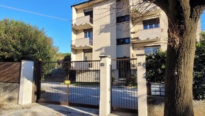 Un immeuble résidentiel à plusieurs étages, doté d’une entrée sécurisée par un portail, avec un arbre au premier plan et un ciel bleu dégagé.