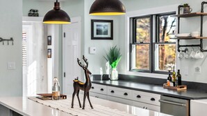 A modern kitchen with a white countertop, two pendant lights, a decorative deer figurine, and a window with a view of trees. - Andover in-town, upscale furnished home, single level living & 2-car garage (Andover)