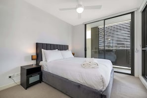 Un dormitorio moderno con cama, mesitas de noche, ventilador de techo y puerta corrediza de vidrio que da a un balcón.