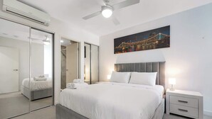 A modern hotel room with a large bed, a ceiling fan, a nightstand, a bedside lamp, a large mirror, and a cityscape painting. - Spacious 2 bedroom Apartment Near PA Hospital (Woolloongabba)