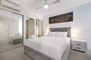 Una moderna camera d'albergo con un grande letto, una ventola a soffitto, un comodino, una lampada da comodino, un ampio specchio e un quadro raffigurante un panorama urbano.