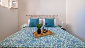 Una cama bien tendida con una colcha estampada, una planta en maceta y un pequeño adorno sobre una bandeja de madera.