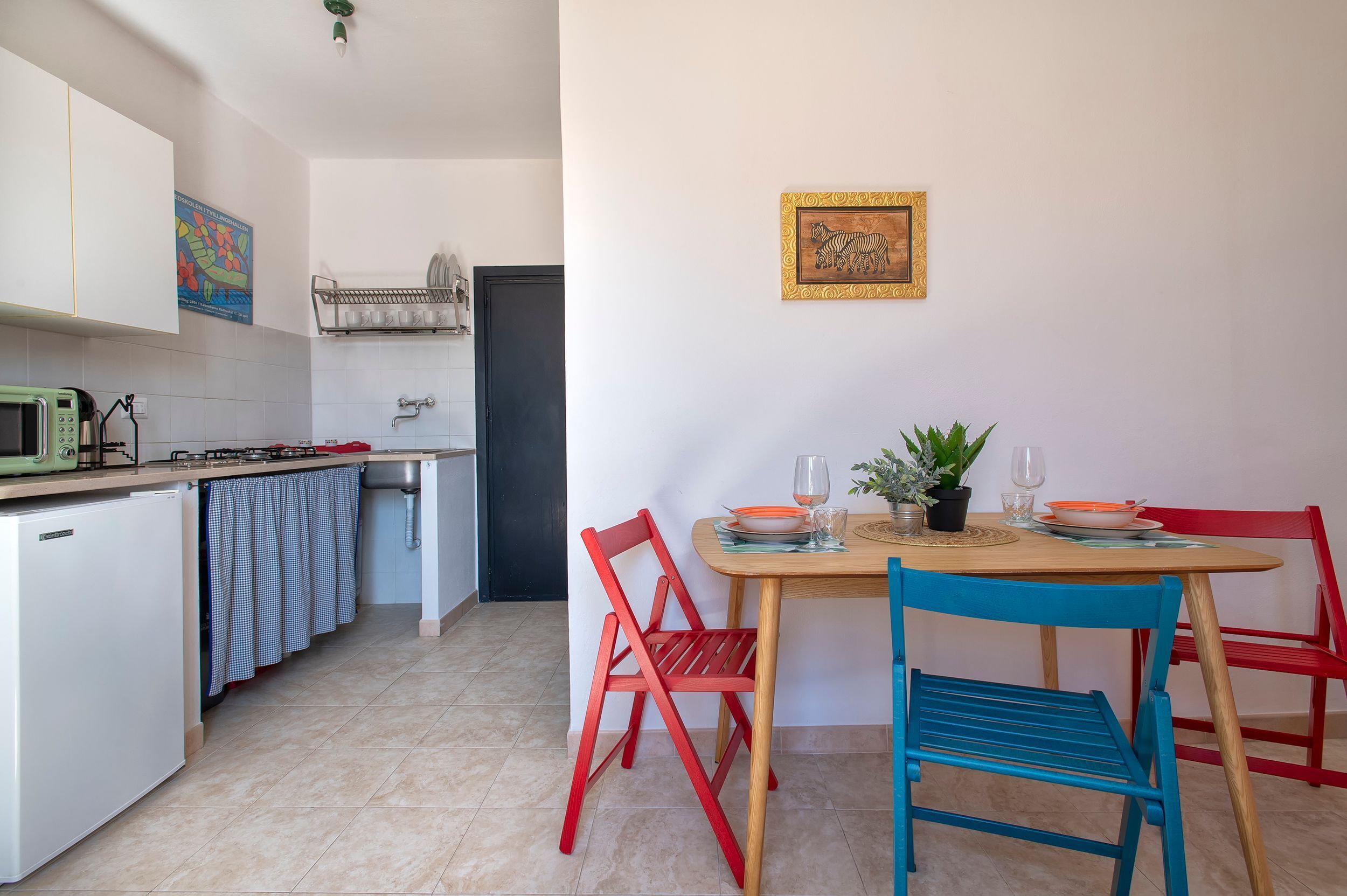A compact kitchen with a microwave, sink, and cabinets. There is a dining table with chairs and a small potted plant.