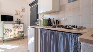 Cocina compacta con microondas, tetera y cocina a gas. Incluye un refrigerador pequeño y una cortina a cuadros.