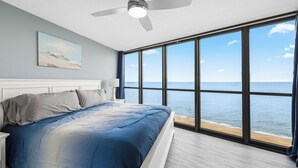 A bedroom with a large window overlooking the ocean, a ceiling fan, and a bed with a blue and white bedding.