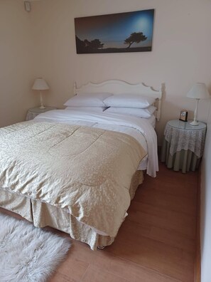 Un letto rifatto con precisione, con testiera bianca, un tappeto bianco soffice e un quadro di un albero al muro.