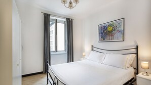 Een slaapkamer met een bed, een nachtkastje, een lamp, een raam met gordijnen en een kunstwerk aan de muur.