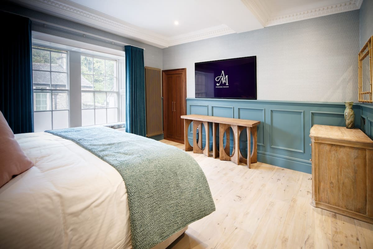 Ein Hotelzimmer mit einem großen Bett, einem an der Wand montierten Fernseher, einer hölzernen Bank und einem Fenster mit Vorhängen.