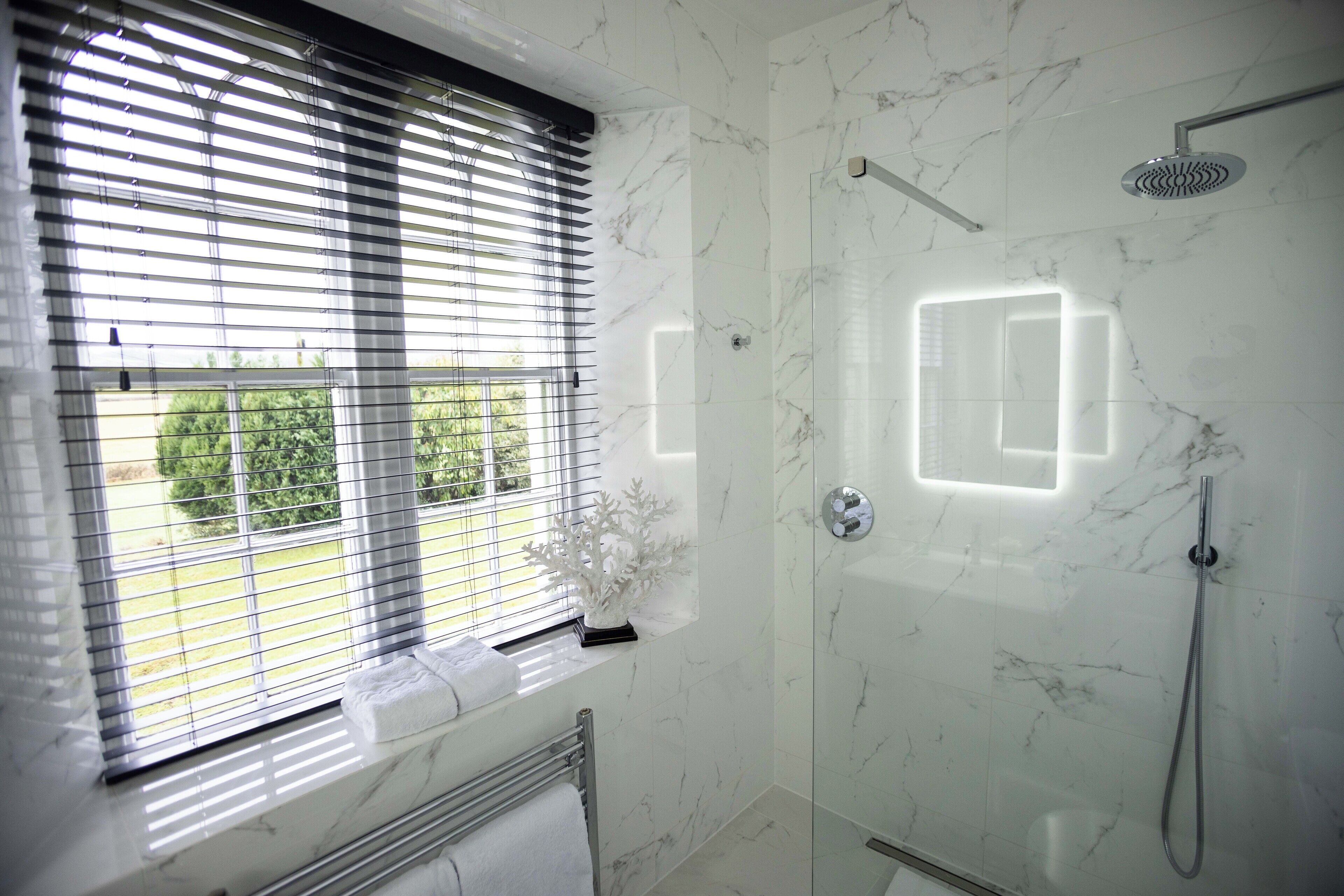 A modern bathroom with a glass-enclosed shower, a window with blinds, and a marble-tiled wall.