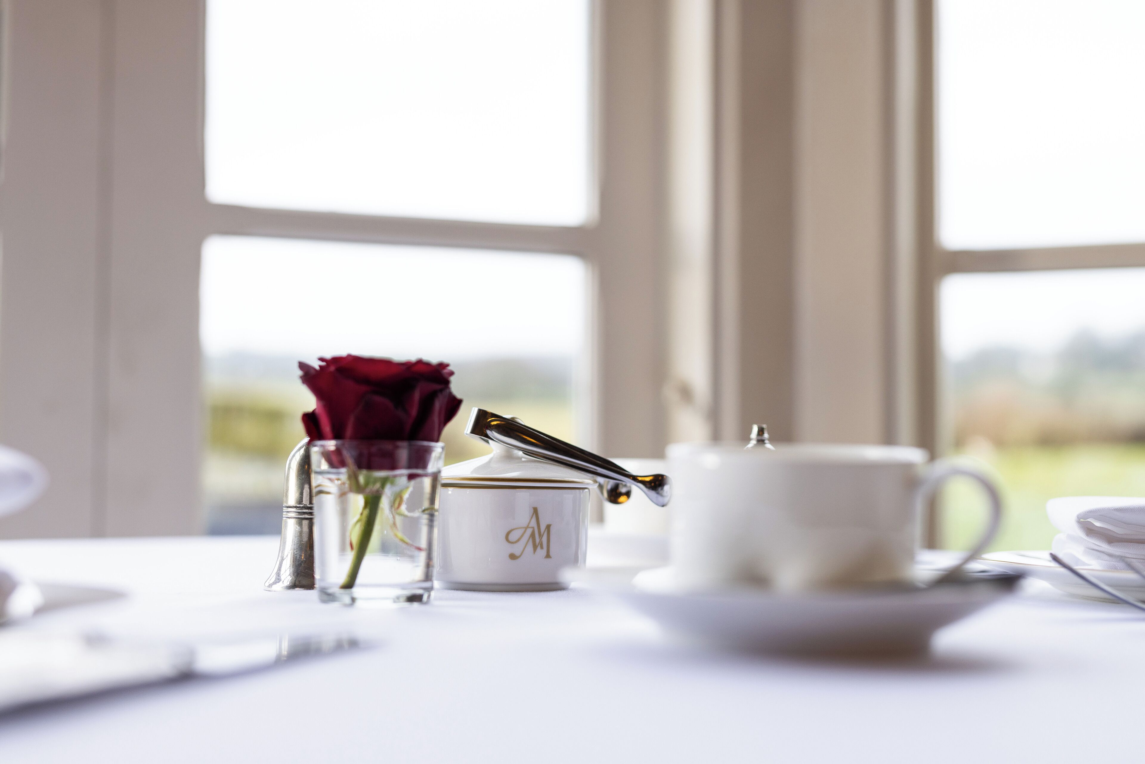 A table with a white teapot, a sugar bowl with a gold emblem, a glass with a red rose, and a clear glass.