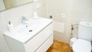 A bathroom with a white sink, faucet, and toilet, and brown tiled flooring. - Asana Surf House (Famara)