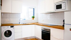 A compact kitchen with white cabinets, a wooden countertop, a washing machine, an oven, and a microwave. - Asana Surf House (Famara)
