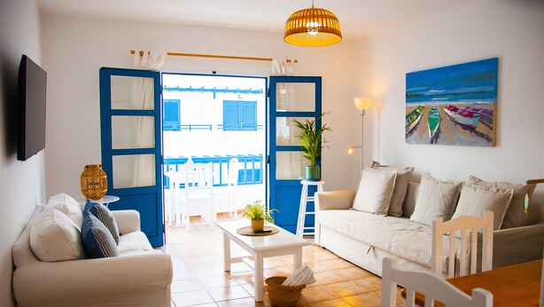 A living room with a white sofa, a white armchair, a wooden table, and a painting of boats on the wall. - Asana Surf House (Famara)