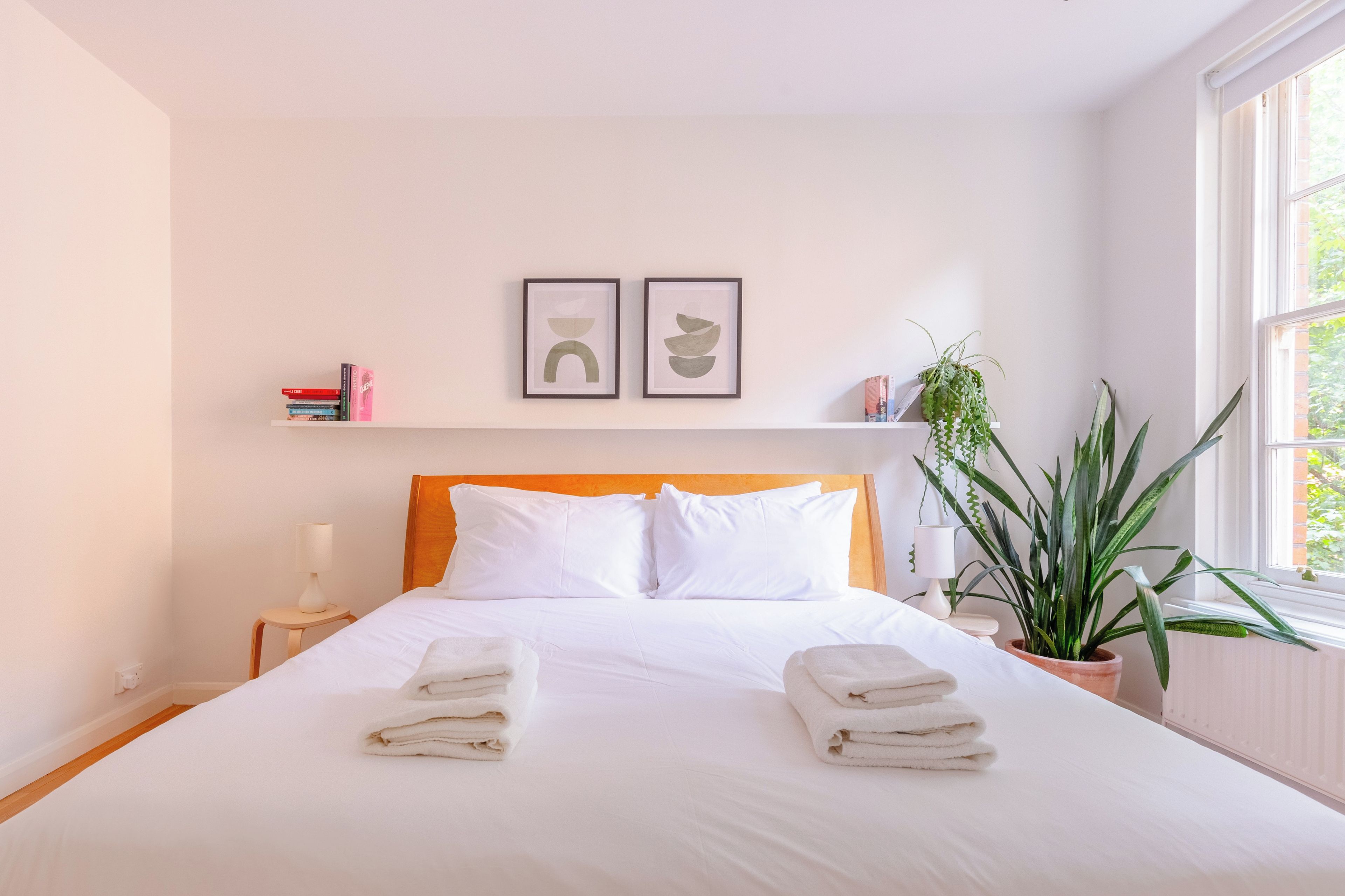 A neatly made bed with white linens and towels, a bedside lamp, a wooden headboard, two framed artworks on the wall, and a potted plant beside the window.