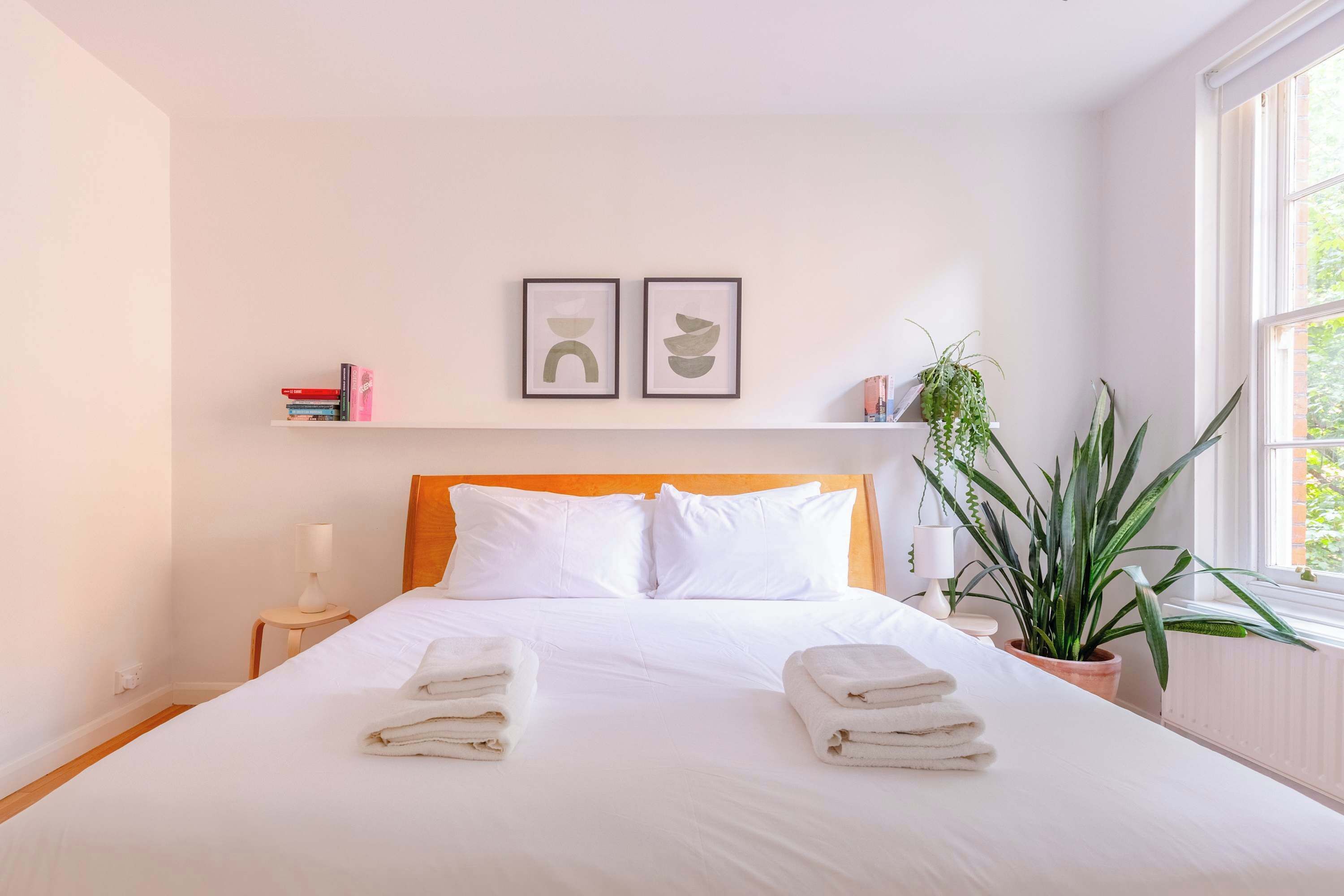 Una cama bien hecha con toallas, una lámpara de noche, un estante con libros y una planta, y una ventana con vistas.
